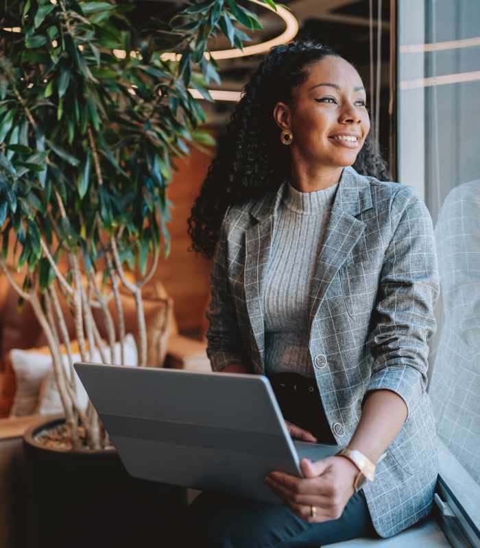 Woman sitting by a window with a laptop, surrounded by greenery and modern decor at Unionville Station Townhomes in Hilton, New York