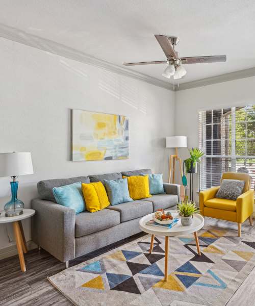 Bright living room with gray sofa, colorful pillows, yellow chair, large windows, and stylish decor at Calder in Round Rock, Texas