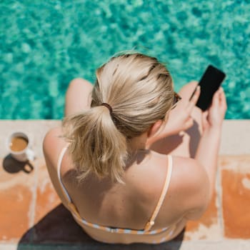 A woman using her phone while she dips her feet in the swimming pool at Cypress Creek at River Bend in Georgetown, Texas