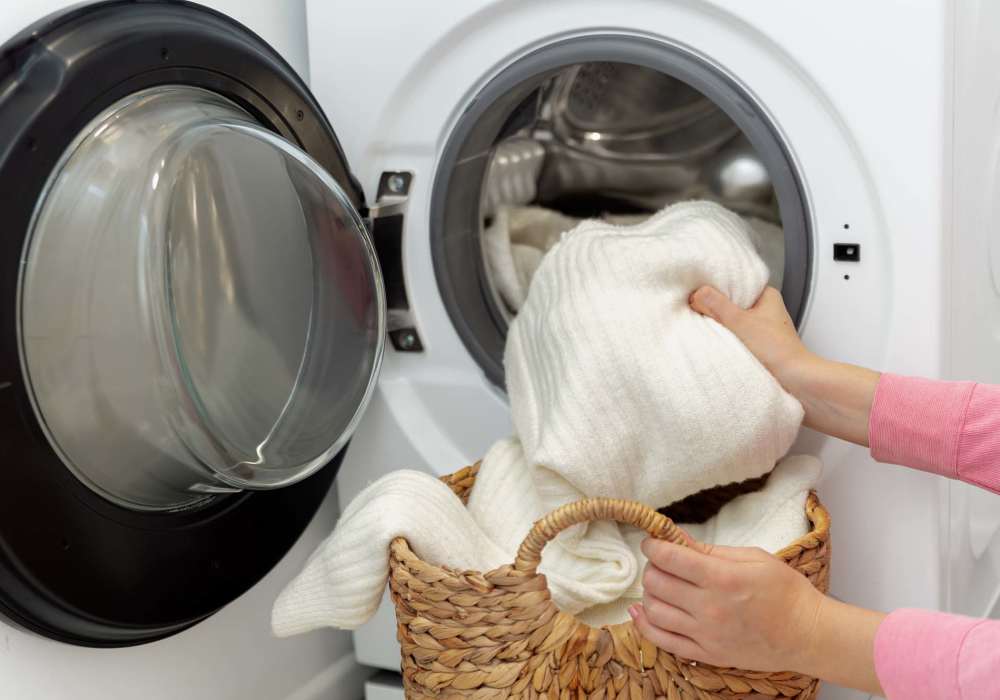 Person placing white blankets into a front-loading washing machine from a woven basket at Magnolia Crossing and Bell Ridge II in Pace, Florida