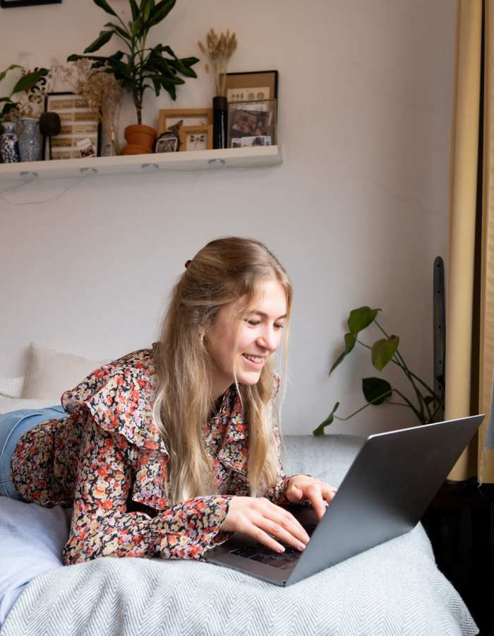 Student working on her laptop while lying on her bed at Midtown 905 in Denton, Texas