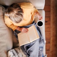 A woman holding a cup of while reading a book on her couch at Residences at Congressional Village in Rockville, Maryland