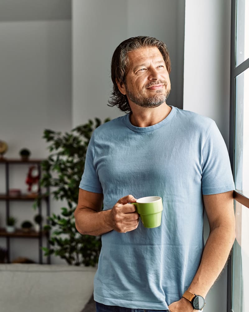 A smiling man holding a cup of coffee and looking out his apartment window at Thirty - One 32 Cypress in Hoover, Alabama