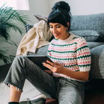 Woman sitting on floor with tablet, cozy living room with plants and a gray sofa at Riverbirch in Ames, Iowa