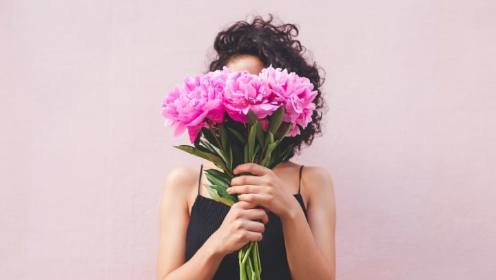 a young woman stands in front of a pale pink wall, her face obscured by a bouquet of pink flowers she hold at eye level | Florists in Gilbert