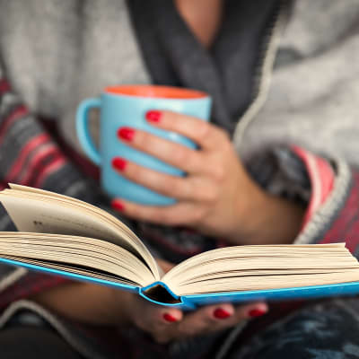 A woman holding a cup of coffee in one hand and reading a book in another at Briarcliff Apartment Homes in Milledgeville, Georgia
