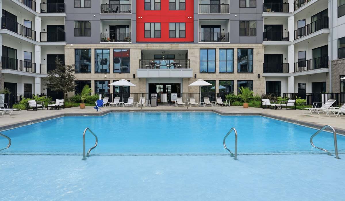 St. Matthews, KY, Pet-Friendly Luxury Apartments - The Station at Middletown - Resort-Style Pool With Lounge Seating, Umbrellas, Apartment Balconies, Large Windows, And Modern Building Exterior.
