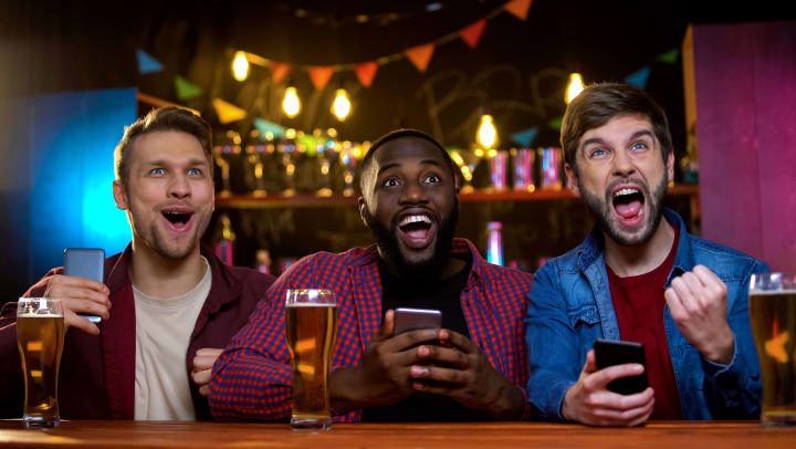 Three men sitting at a bar with beers, cheering.