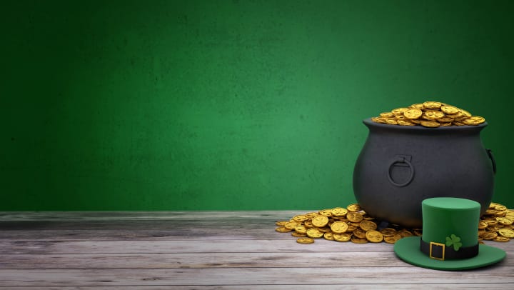 Pot of gold, a pile of gold coins, and green leprechaun hat sitting on a wooden floor with a green wall in the background