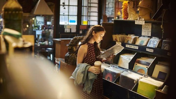 A woman in a brown dress with white polka dots holds a vinyl record while in an antique store, which also shows background items such as sculptures, lamps, and glass cases full of items