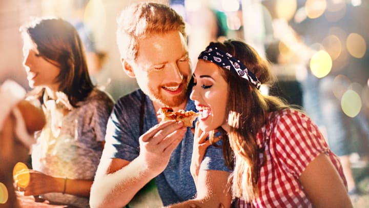  Man and a woman are sitting at a table with friends. Man is feeding the woman a slice of pizza. 