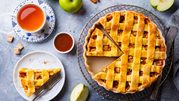  Apple pie on a cooling rack, on a table, with a slice cut out, sitting next to various ingredients and a cup of tea. 