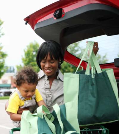 Woman unloading grocery bags from a car trunk while holding a toddler in a parking lot at Cypress Run in Orlando, Florida