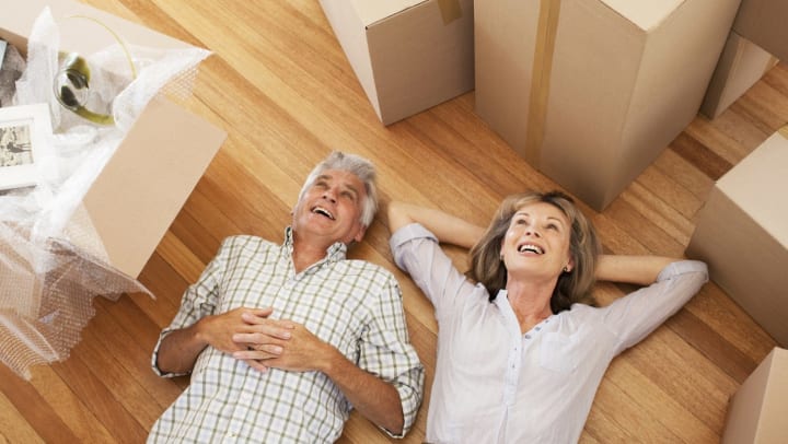 Older man and older woman smiling and lying on hardwood floor surrounded by cardboard boxes