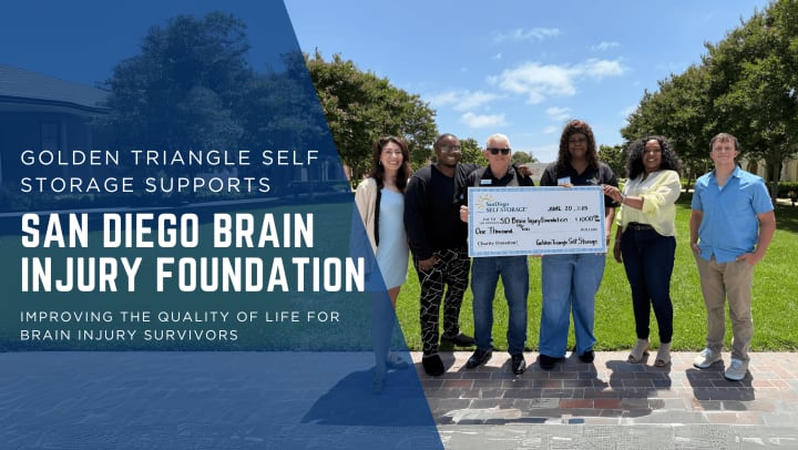 Golden Triangle Self Storage team presents a $1,000 donation check to the San Diego Brain Injury Foundation at Liberty Station in Point Loma. Six people stand smiling outdoors on a sunny day, supporting programs that improve quality of life for brain injury survivors.