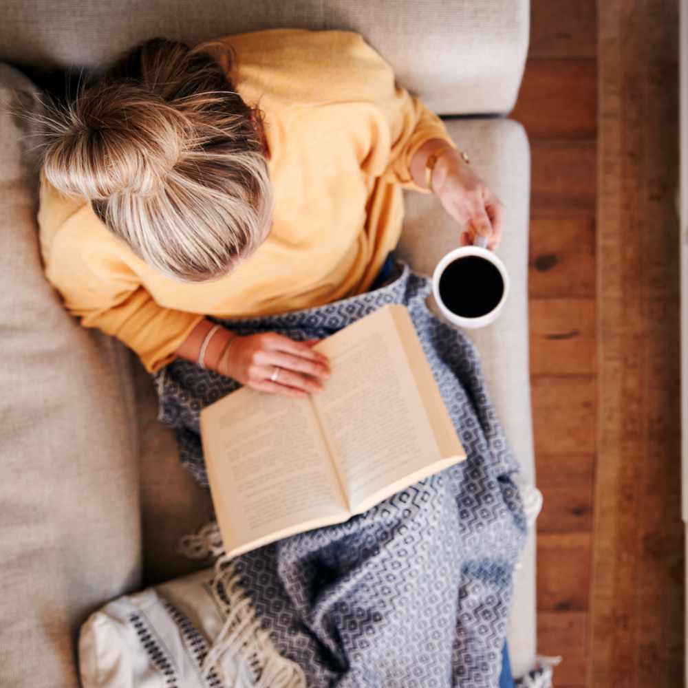 Resident woman reading a book and holding a cup of coffee while sitting on a couch at Sycamore Villas in Nashville, Tennessee