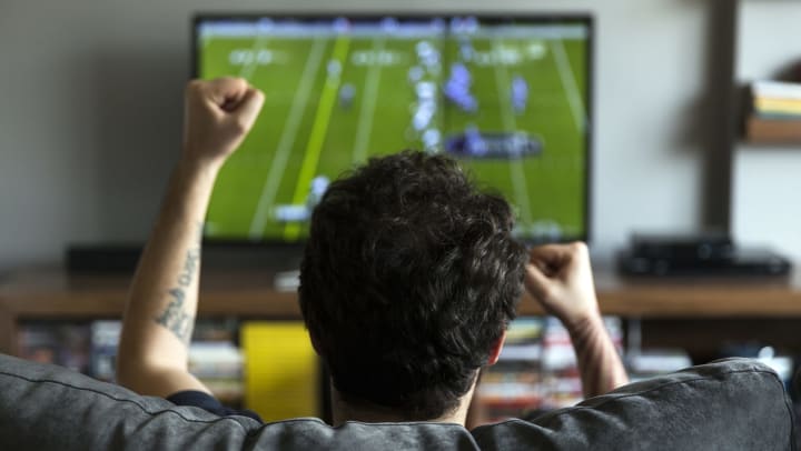 A man in the foreground cheering. In the background is a TV playing a football game. 