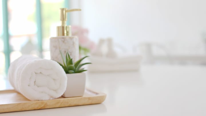A white bathroom counter with a wooden tray holding a rolled white towel, a succulent plant in a white pot, and a brass and marble soap dispenser. 