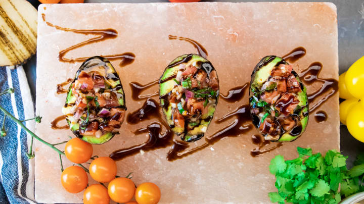 Three avocados filled with Bruschetta filling on a slab of himalayan salt, with cherry tomatoes, charred onion, bell peppers, and a bunch of cilantro