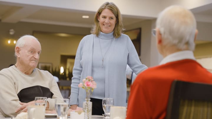 A smiling culinary director stands and engages warmly with two seated residents at a dining table, reflecting Clearwater’s personalized and attentive dining approach.